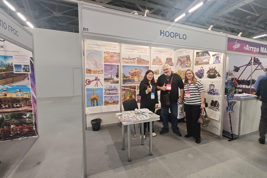 Команда Hooplo Rides позирует с парой посетителей на стенде B5 выставки RAAPA EXPO Осень 2026 в Москве, на досках портфолио представлены аттракционы для парков развлечений и развлекательное оборудование.
