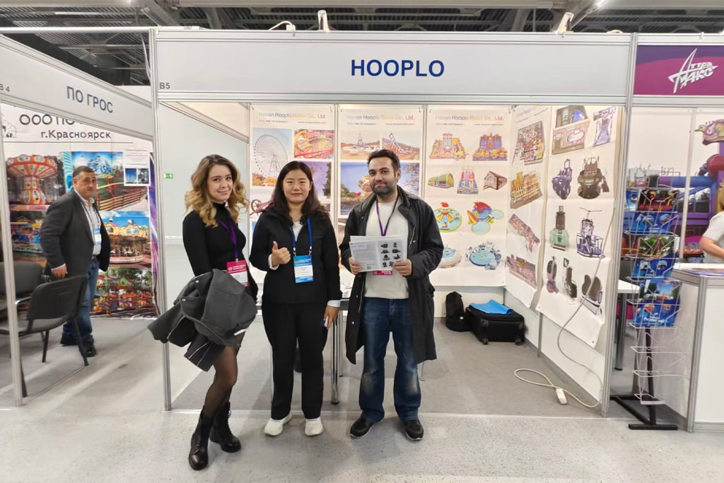 Сотрудница Hooplo Rides позирует с новыми клиентами на стенде B5 выставки RAAPA EXPO Осень 2026 в Москве, на стене стенда размещены постеры и каталоги аттракционов.