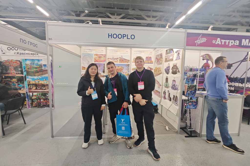 Представительница Hooplo Rides делает групповое фото с двумя посетителями-мужчинами на стенде Hooplo, RAAPA EXPO Осень 2026 Москва, за спиной постеры аттракционов и паркового развлекательного оборудования.
